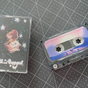 Può includere: Due musicassette trasparenti. Una ha una copertina nera con un'illustrazione di un cartone animato di una ragazza e un cane, con la scritta "BEE and Puppycat". L'altra cassetta ha un design sfumato rosa, bianco e blu.