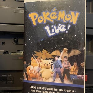 May include: A VHS tape for "Pokémon Live!" features a starry background with the title in yellow and blue. The cover shows costumed characters and performers. The text at the bottom lists the cast and crew.