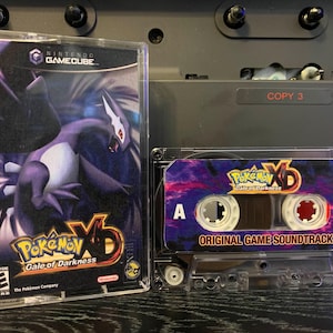 May include: A Nintendo GameCube game case and a cassette tape. The game case features the "Pokémon XD: Gale of Darkness" title and artwork of a purple dragon. The cassette tape is labeled "Original Game Soundtrack".