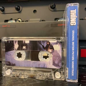 May include: A clear cassette tape of "The Thing" original motion picture soundtrack. The cassette case shows a scene from the movie. The spine of the cassette case is blue with white text that reads "THING" and "ORIGINAL MOTION PICTURE SOUNDTRACK".