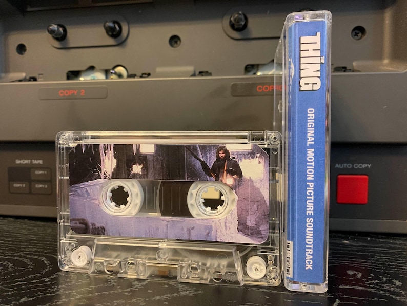 May include: A clear cassette tape of "The Thing" original motion picture soundtrack. The cassette case shows a scene from the movie. The spine of the cassette case is blue with white text that reads "THING" and "ORIGINAL MOTION PICTURE SOUNDTRACK".