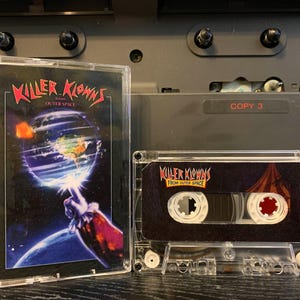 May include: Two clear cassette tapes with the text "KILLER KLOWNS FROM OUTER SPACE". The cassette cases have a dark background with a colorful image of a clown's hand pointing at a planet. The tapes are displayed in front of a vintage audio device.