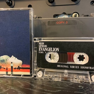 May include: Two clear cassette tapes. One has a blue cover with a person's face. The other has a black label with the text "NEON GENESIS EVANGELION" and "ORIGINAL SERIES SOUNDTRACK".