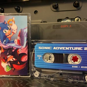 Peut inclure: Deux cassettes audio avec Sonic le Hérisson et Shadow le Hérisson. L'une présente une image colorée de Sonic et Shadow. L'autre est étiquetée "SONIC ADVENTURE 2" avec un design bleu et blanc.