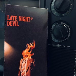 May include: A VHS tape with the title "LATE NIGHT with the DEVIL" in red text. The cover art features a person in a suit with a flaming head. Beside the tape is a black vintage General Electric television with dials.