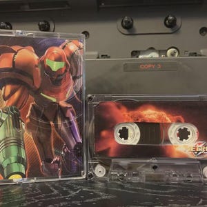 Peut inclure: Deux cassettes transparentes. L'une présente une illustration colorée d'un personnage en armure orange avec une visière verte. L'autre cassette a un design orange et rouge flamboyant avec le mot "METROID".