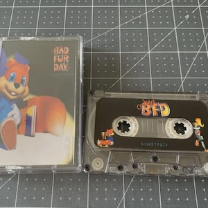 Könnte beinhalten: Eine transparente Kassette und eine Kassette werden gezeigt. Das Kassettenetui zeigt eine Cartoonfigur in Blau und Orange. Auf der Kassette steht "BFD" und "Soundtrack".