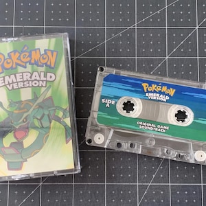 Puede incluir: Una cinta de cassette transparente con una etiqueta verde y azul con el texto "Pokémon Emerald Version" y un gráfico de una criatura con forma de dragón. La caja de la cinta de cassette está al lado de la cinta.