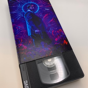 May include: A black JVC VHS tape with a colorful movie cover featuring a man in a suit holding a gun. The cover has a purple and blue background with a halo around the man's head.