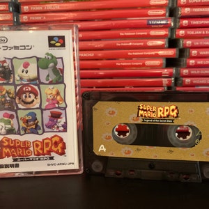 Puede incluir: Una cinta de casete y una caja de juego de Super Mario RPG. La caja del juego presenta un collage colorido de personajes, incluyendo a Mario, Yoshi y la Princesa Peach. La cinta de casete tiene el título del juego y un diseño floral.