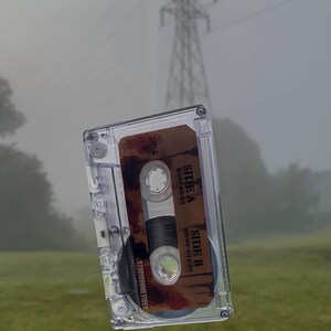 May include: A clear cassette tape with a brown label featuring the text "SIDE A" and "SIDE B" with time stamps. The cassette is set against a blurred background of a grassy field and a power line.