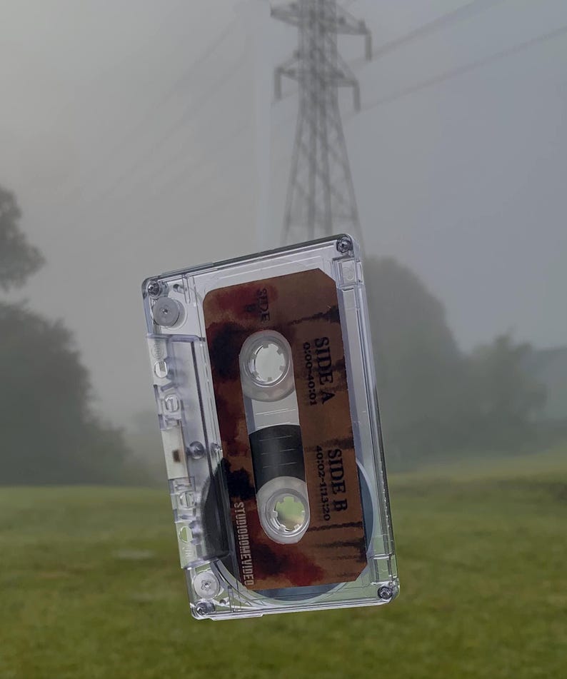 May include: A clear cassette tape with a brown label featuring the text "SIDE A" and "SIDE B" with time stamps. The cassette is set against a blurred background of a grassy field and a power line.