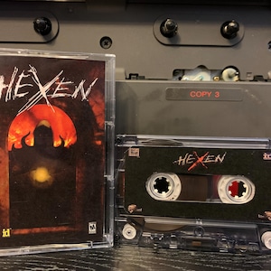 Puede incluir: Una cinta de casete transparente y su estuche para el videojuego "Hexen". La carátula presenta el título del juego en blanco, con un gráfico naranja y marrón llameante. La cinta de casete es negra con texto blanco.