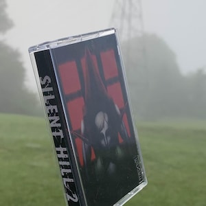 May include: A clear cassette tape with a black and red cover featuring the text "SILENT HILL 2". The cover art depicts a dark figure with a red hood. The cassette is set against a blurred green and gray background.