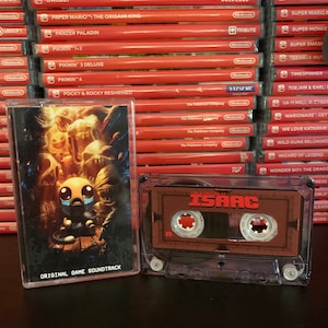 Puede incluir: Una cinta de cassette transparente con una etiqueta marrón con el texto "THE BINDING OF ISAAC" y un título rojo. La cinta de cassette está junto a una caja de juego con el texto "ORIGINAL GAME SOUNDTRACK". Una pila de cajas de juegos rojas de Nintendo Switch está en el fondo.