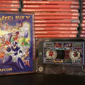 Puede incluir: Un cartucho de videojuego y una cinta de casete de Mega Man X. El cartucho tiene una colorida ilustración de personajes con el logotipo de Capcom. La cinta de casete es transparente con un gráfico colorido. Hay cartuchos de juego rojos en el fondo.