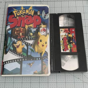 May include: A VHS tape and a Nintendo 64 game cartridge for Pokémon Snap. The VHS tape has a colorful cover featuring Pikachu and other Pokémon characters. The cartridge is black with a smaller image of the game's cover art.
