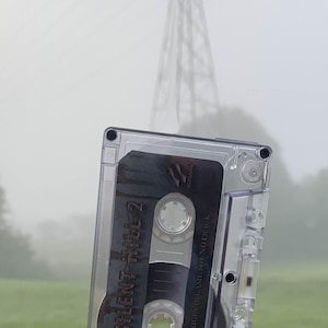 May include: A clear cassette tape with the text "Silent Hill 2 Original Game Soundtrack" floats in front of a blurred background of a power line and a green field. The tape is in a clear plastic case.