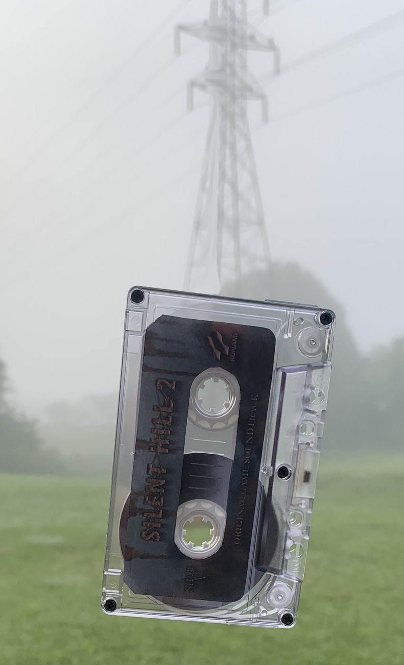 May include: A clear cassette tape with the text "Silent Hill 2 Original Game Soundtrack" floats in front of a blurred background of a power line and a green field. The tape is in a clear plastic case.
