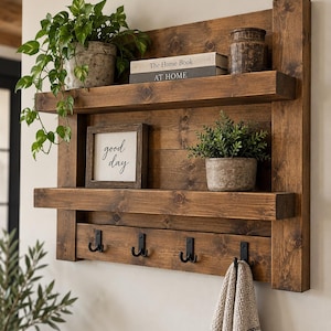 May include: A wall-mounted wooden shelf unit with two horizontal shelves and four black metal double hooks. The unit features a dark brown wood grain finish. Items displayed include two potted green plants, two books, and a framed sign reading good day.