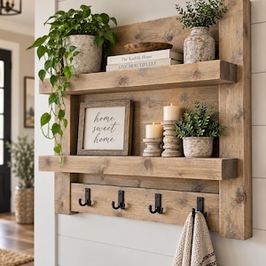 May include: A wall-mounted wooden shelf unit with two horizontal shelves and four black metal hooks. The unit features a framed sign reading home sweet home, two pillar candles, two potted plants, a wooden bowl, and two stacked books.