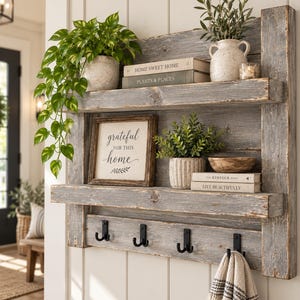 May include: A wall-mounted rustic gray wooden shelf unit featuring two horizontal shelves and four black metal double hooks. The shelves hold potted green plants, a framed sign reading grateful for this home, and books titled Home Sweet Home, Plants & Places, The Kinfolk Home, and Live Beautifully.