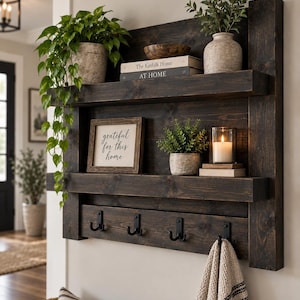 May include: A dark stained wooden wall shelf featuring two horizontal shelves and four black metal hooks. The shelves hold a trailing green plant, two books titled The Kinfolk Home and AT HOME, a small wooden bowl, a framed sign reading grateful for this home, a potted plant, and a white candle in a glass holder.