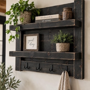 May include: A wall-mounted black wooden shelf unit with two horizontal shelves and four black metal hooks at the bottom. The unit features a distressed finish and holds a potted plant, two books titled The Home Book and AT HOME, a small framed sign reading good day, and a second potted plant.