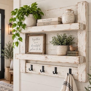 May include: A wall-mounted white distressed wood shelf unit featuring two horizontal shelves and a bottom row with four black metal double hooks. The shelves hold a potted plant, two books labeled Live Simply and Grateful, a vase, and a framed sign.