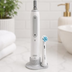 Puede incluir: Un cepillo de dientes eléctrico blanco y gris con un cabezal de repuesto. El cepillo de dientes tiene un botón de encendido y un símbolo de Bluetooth. El cabezal del cepillo tiene cerdas azules y blancas. El cepillo de dientes está en una base gris moteada.