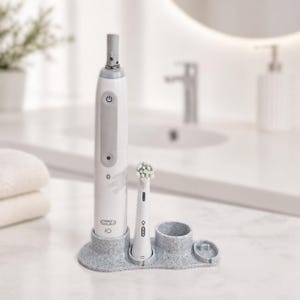 May include: A white and grey Oral-B iO electric toothbrush with a charging base and an extra brush head. The toothbrush has a power button and a Bluetooth symbol. The base is speckled grey and has a holder for the brush and an extra head.