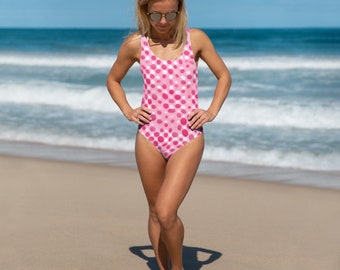 May include: A pink and white polka dot swimsuit with a scoop neck and thin straps. The swimsuit is on a woman standing on a sandy beach.