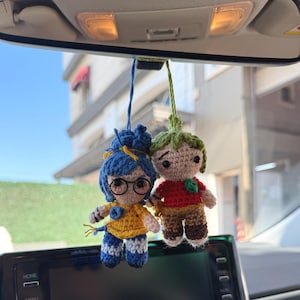 Pode incluir: Dois amuletos de carro de croch&ecirc; pendurados no espelho retrovisor de um carro. Um amuleto &eacute; uma menina com cabelo azul, &oacute;culos e uma blusa amarela. O outro &eacute; um menino com cabelo verde e uma camisa vermelha. O painel do carro e um edif&iacute;cio s&atilde;o vis&iacute;veis.