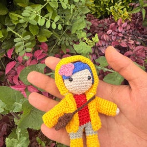 May include: A small crocheted doll featuring blue hair with a pink flower accessory, wearing a bright yellow hooded raincoat over a red shirt and gray pants, with yellow boots and a small brown cross-body bag, crafted with detailed yarn stitching.