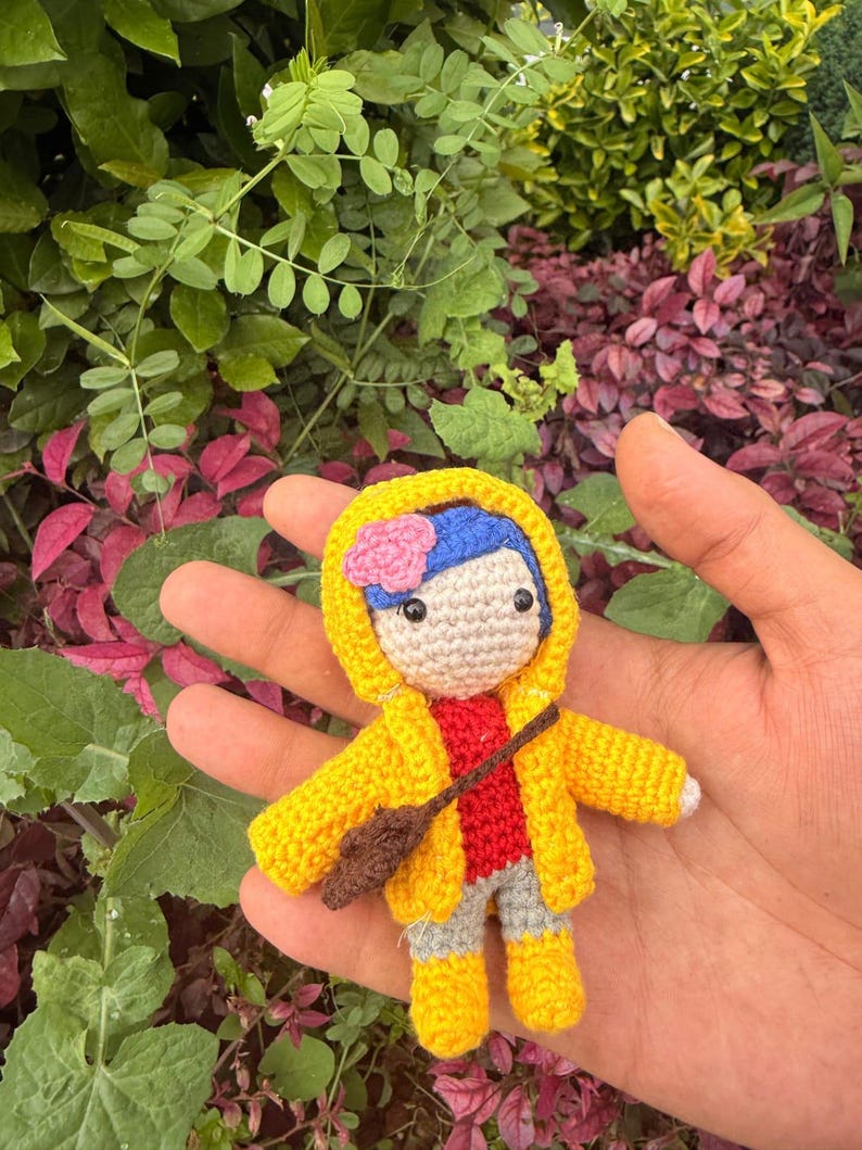 May include: A small crocheted doll featuring blue hair with a pink flower accessory, wearing a bright yellow hooded raincoat over a red shirt and gray pants, with yellow boots and a small brown cross-body bag, crafted with detailed yarn stitching.
