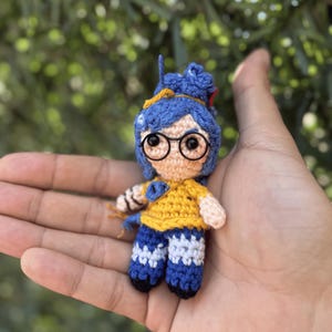 Pode incluir: Pequena boneca de croch&ecirc; feita &agrave; m&atilde;o, com cabelo azul, &oacute;culos redondos e uma camisa amarela. A boneca est&aacute; vestindo cal&ccedil;as azuis com detalhes brancos. A boneca &eacute; segurada na m&atilde;o de uma pessoa contra um fundo verde desfocado.