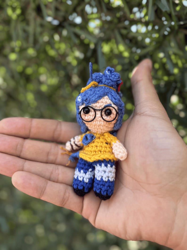 Pode incluir: Pequena boneca de croch&ecirc; feita &agrave; m&atilde;o, com cabelo azul, &oacute;culos redondos e uma camisa amarela. A boneca est&aacute; vestindo cal&ccedil;as azuis com detalhes brancos. A boneca &eacute; segurada na m&atilde;o de uma pessoa contra um fundo verde desfocado.