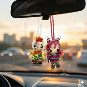 Op de afbeelding: Twee handgemaakte gehaakte poppen die aan de achteruitkijkspiegel van een auto hangen. De poppen hebben gedetailleerde kenmerken, waaronder verschillende gekleurde outfits en kapsels. De ene pop heeft oranje haar, de andere roze. De achtergrond toont een vage stadsgezicht.