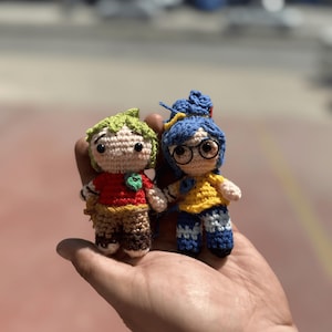 Pode incluir: Duas bonecas de croch&ecirc; feitas &agrave; m&atilde;o. Uma boneca tem cabelo verde, uma camisa vermelha e cal&ccedil;as marrons. A outra boneca tem cabelo azul, uma camisa amarela e cal&ccedil;as azuis. Ambas as bonecas t&ecirc;m olhos pretos e s&atilde;o seguradas em uma m&atilde;o.