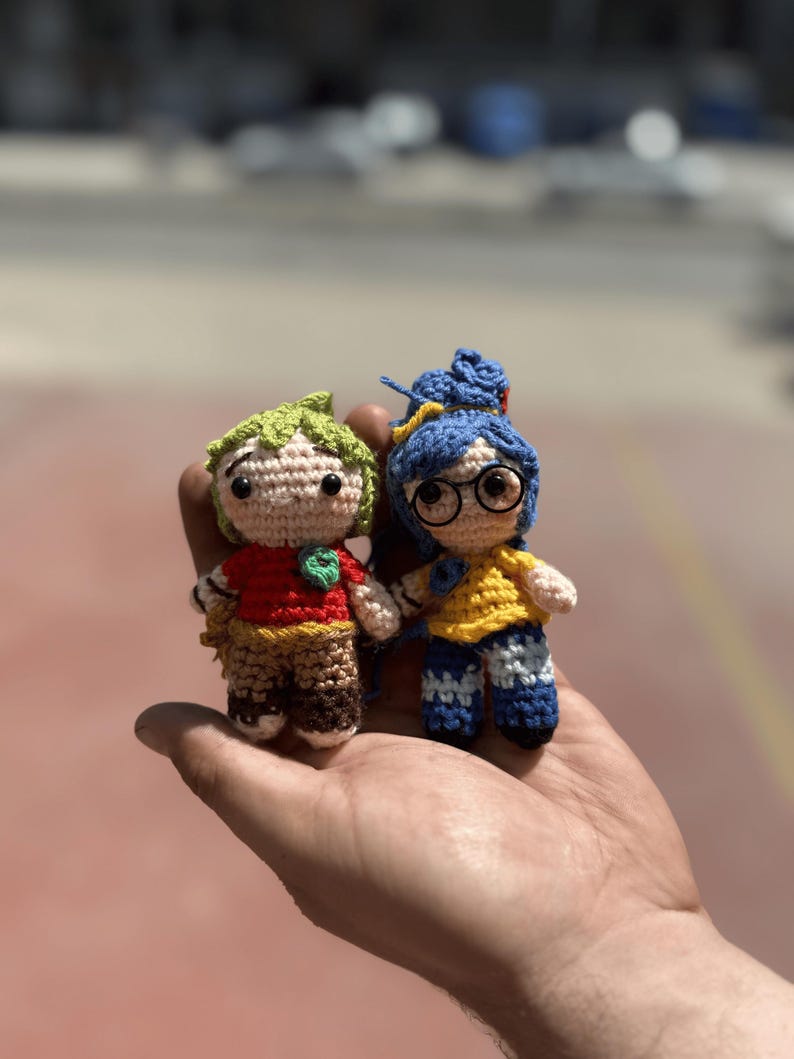 Pode incluir: Duas bonecas de croch&ecirc; feitas &agrave; m&atilde;o. Uma boneca tem cabelo verde, uma camisa vermelha e cal&ccedil;as marrons. A outra boneca tem cabelo azul, uma camisa amarela e cal&ccedil;as azuis. Ambas as bonecas t&ecirc;m olhos pretos e s&atilde;o seguradas em uma m&atilde;o.