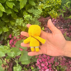 May include: A small, handmade crochet doll featuring a bright yellow hooded jacket, a brown crossbody strap, and light gray legs. The doll is crafted with a textured yarn stitch pattern, showcasing a rounded head, outstretched arms, and short legs.