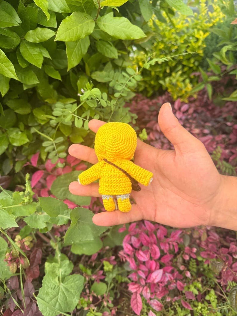 May include: A small, handmade crochet doll featuring a bright yellow hooded jacket, a brown crossbody strap, and light gray legs. The doll is crafted with a textured yarn stitch pattern, showcasing a rounded head, outstretched arms, and short legs.