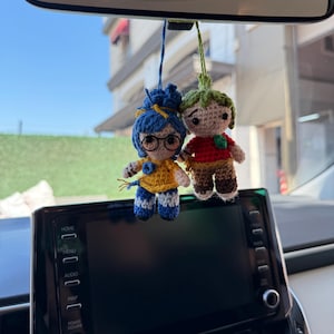 Pode incluir: Duas bonecas de croch&ecirc; feitas &agrave; m&atilde;o penduradas no espelho retrovisor de um carro. Uma boneca tem cabelo azul, &oacute;culos e uma blusa amarela. A outra boneca tem cabelo verde e uma camisa vermelha. As bonecas s&atilde;o presas por fios azuis e verdes.
