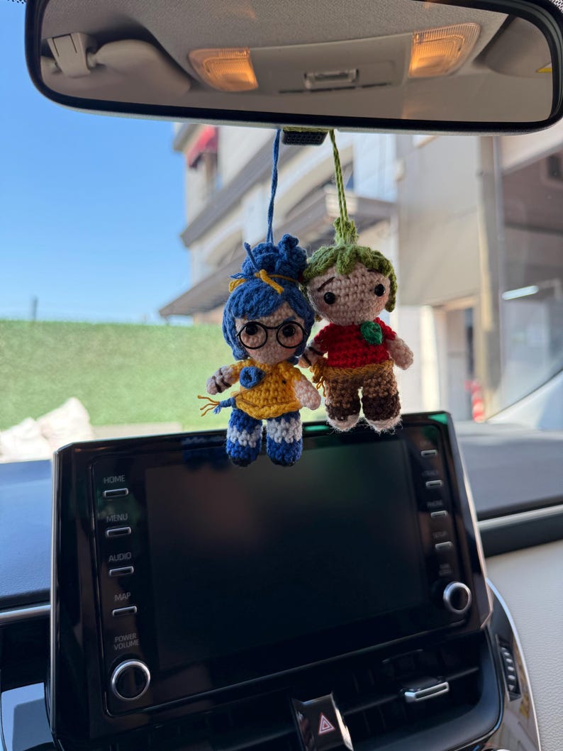 Pode incluir: Duas bonecas de croch&ecirc; feitas &agrave; m&atilde;o penduradas no espelho retrovisor de um carro. Uma boneca tem cabelo azul, &oacute;culos e uma blusa amarela. A outra boneca tem cabelo verde e uma camisa vermelha. As bonecas s&atilde;o presas por fios azuis e verdes.