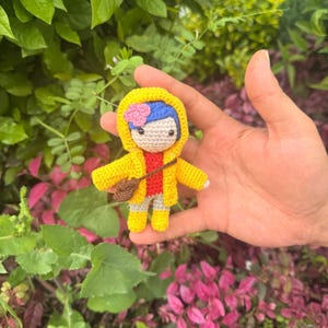 May include: A small crochet doll featuring blue hair with a pink flower, wearing a bright yellow hooded raincoat over a red shirt and gray pants. The doll has black button eyes and carries a small brown cross-body bag, with yellow boots.