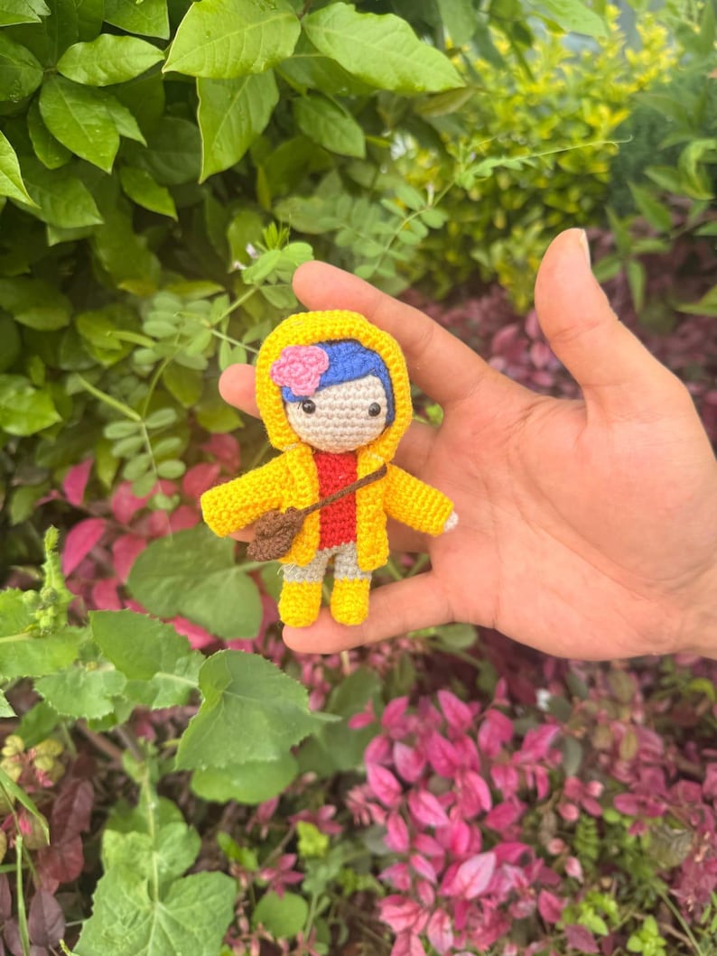 May include: A small crochet doll featuring blue hair with a pink flower, wearing a bright yellow hooded raincoat over a red shirt and gray pants. The doll has black button eyes and carries a small brown cross-body bag, with yellow boots.