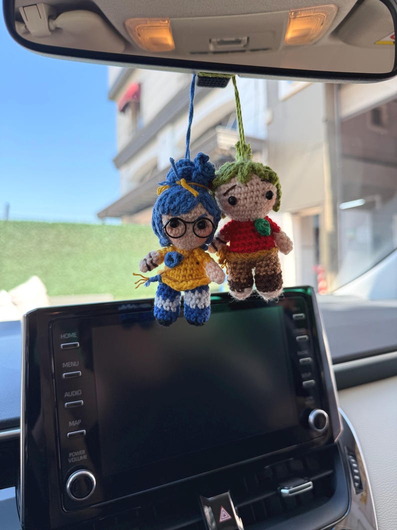 Pode incluir: Dois amuletos de carro de croch&ecirc; pendurados no espelho retrovisor de um carro. Um amuleto &eacute; uma menina com cabelo azul, &oacute;culos e uma blusa amarela. O outro &eacute; um menino com cabelo verde e uma camisa vermelha. O painel do carro e um edif&iacute;cio s&atilde;o vis&iacute;veis.