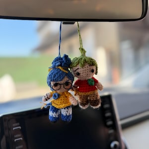 Pode incluir: Duas bonecas de croch&ecirc; penduradas no espelho retrovisor de um carro. Uma boneca tem cabelo azul, &oacute;culos e uma blusa amarela. A outra boneca tem uma cabe&ccedil;a verde e uma camisa vermelha. Ambas as bonecas t&ecirc;m detalhes em castanho e azul.