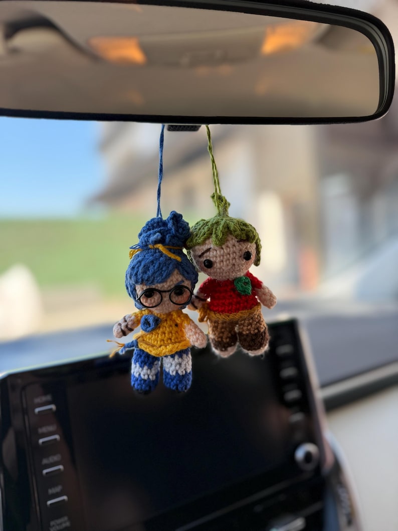 Pode incluir: Duas bonecas de croch&ecirc; penduradas no espelho retrovisor de um carro. Uma boneca tem cabelo azul, &oacute;culos e uma blusa amarela. A outra boneca tem uma cabe&ccedil;a verde e uma camisa vermelha. Ambas as bonecas t&ecirc;m detalhes em castanho e azul.