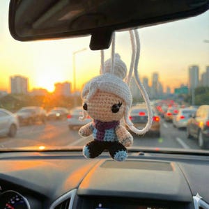 Könnte beinhalten: Ein handgefertigter Häkelpuppen-Autoanhänger. Die Puppe hat einen weißen Hut und einen Schal mit lila, blauen und weißen Streifen. Sie hängt an einem Rückspiegel in einem Auto, mit einer Skyline und Verkehr im Hintergrund.